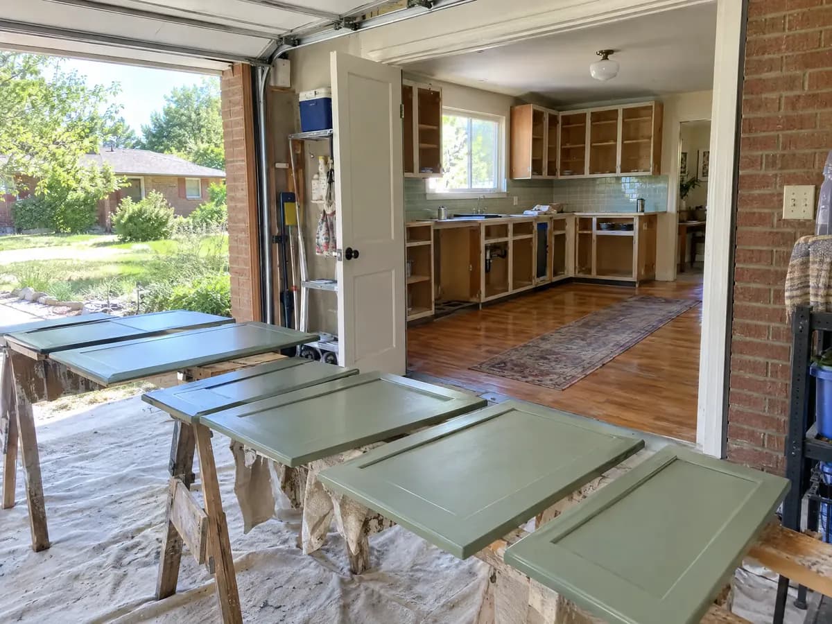 Sage green cabinet doors drying on sawhorses in a garage, with the kitchen's bare cabinet boxes still on the wall beyond