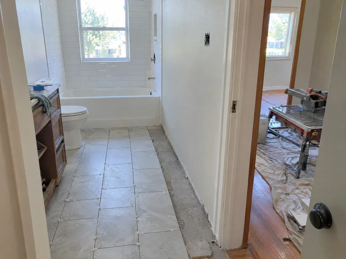 A long narrow hall bathroom with new floor tile half laid from the tub toward the door, spacers in the joints