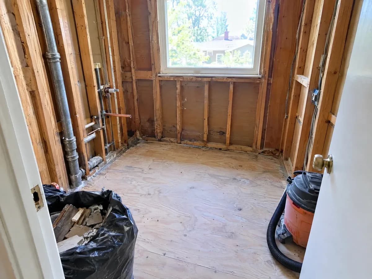 A small hall bathroom gutted to the studs: the tub gone, old pipes in the wall, the subfloor swept, a bag of debris in the doorway