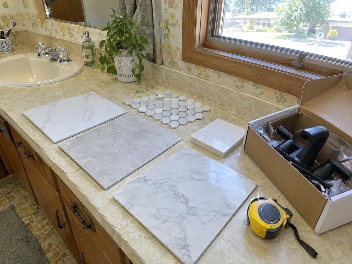 Bathroom tile samples, a hexagon mosaic sheet, a quartz chip, a boxed black faucet and a tape measure on an old vanity