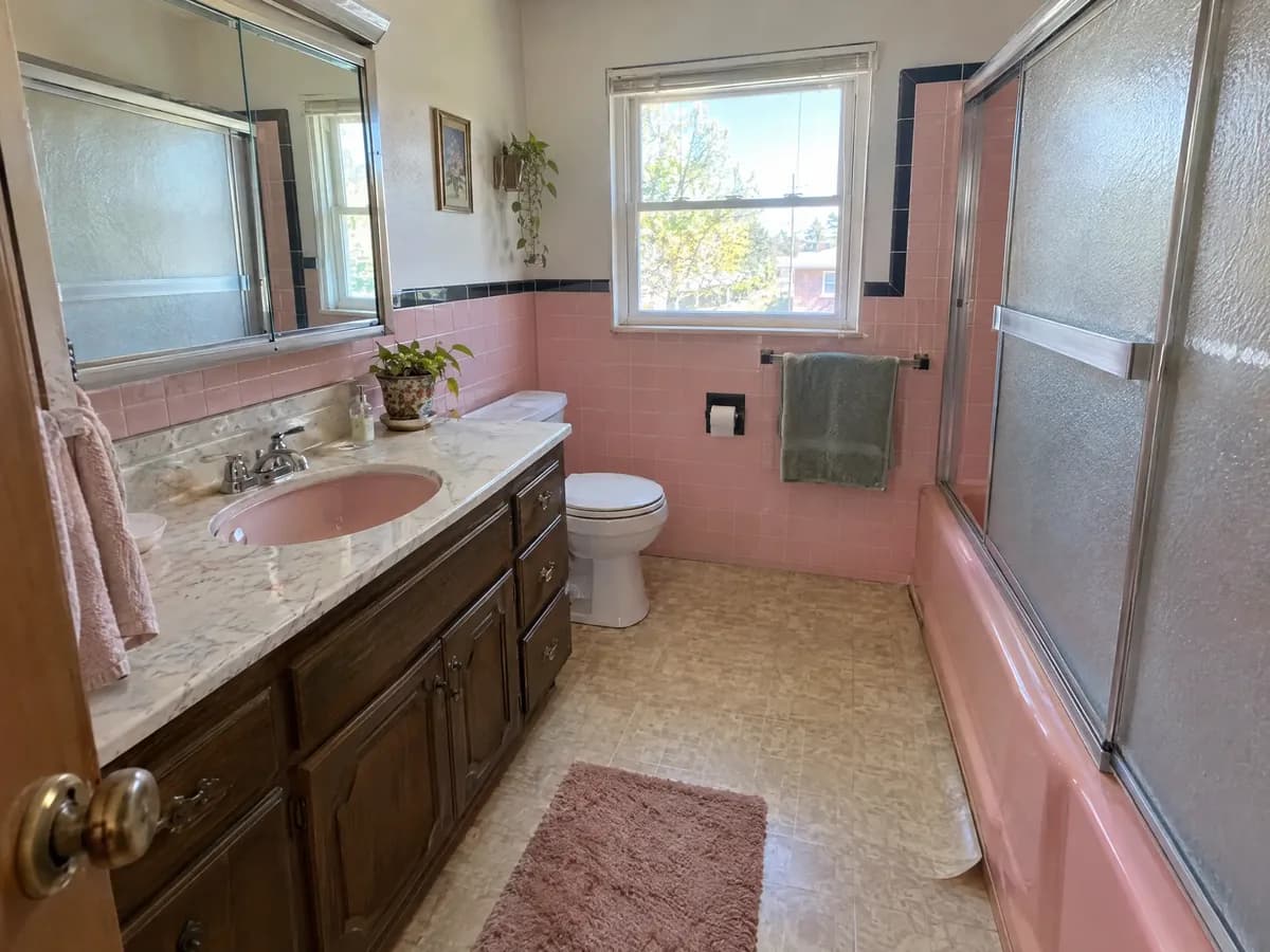 An original 1960s bathroom with pink wall tile, a pink tub behind frosted glass doors and a dark wood vanity