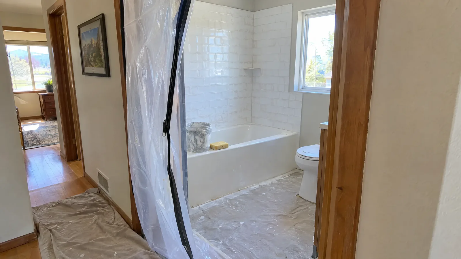 A hall bathroom mid-remodel behind a zippered plastic dust wall, new white tile on the tub surround with grout haze still on it