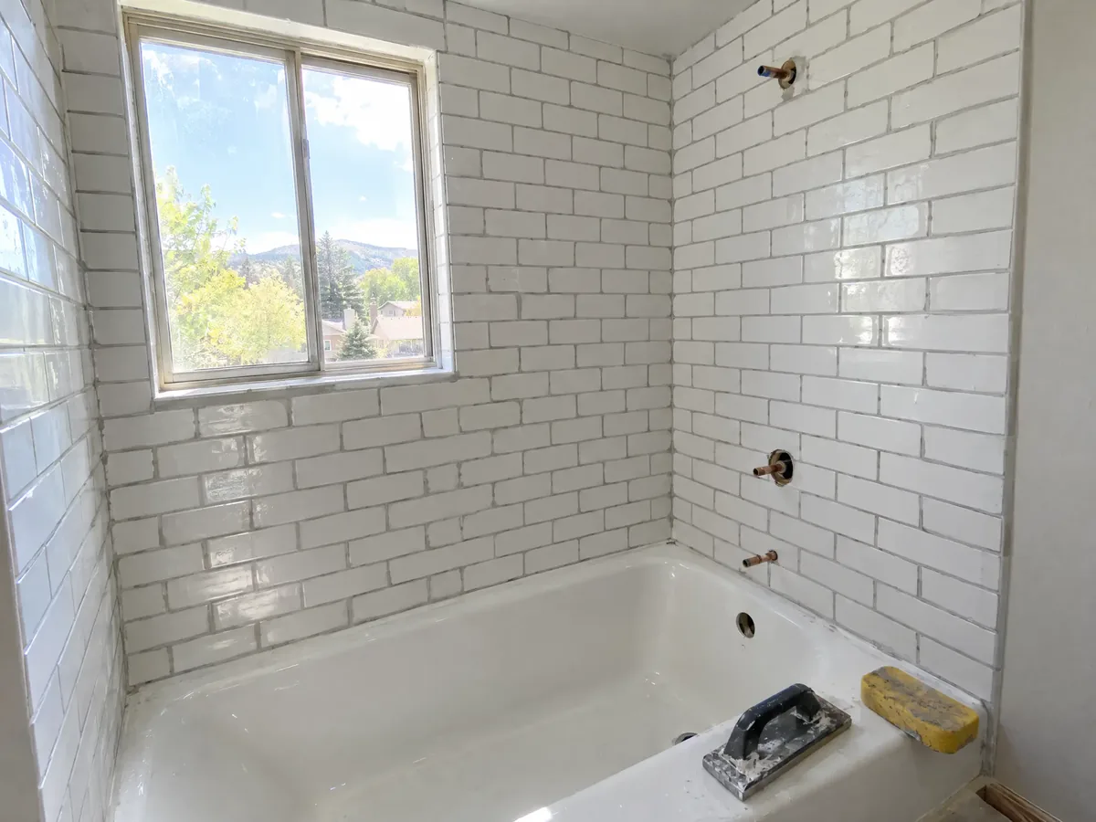 Freshly grouted white subway tile on a tub surround, a grout float and sponge on the tub edge