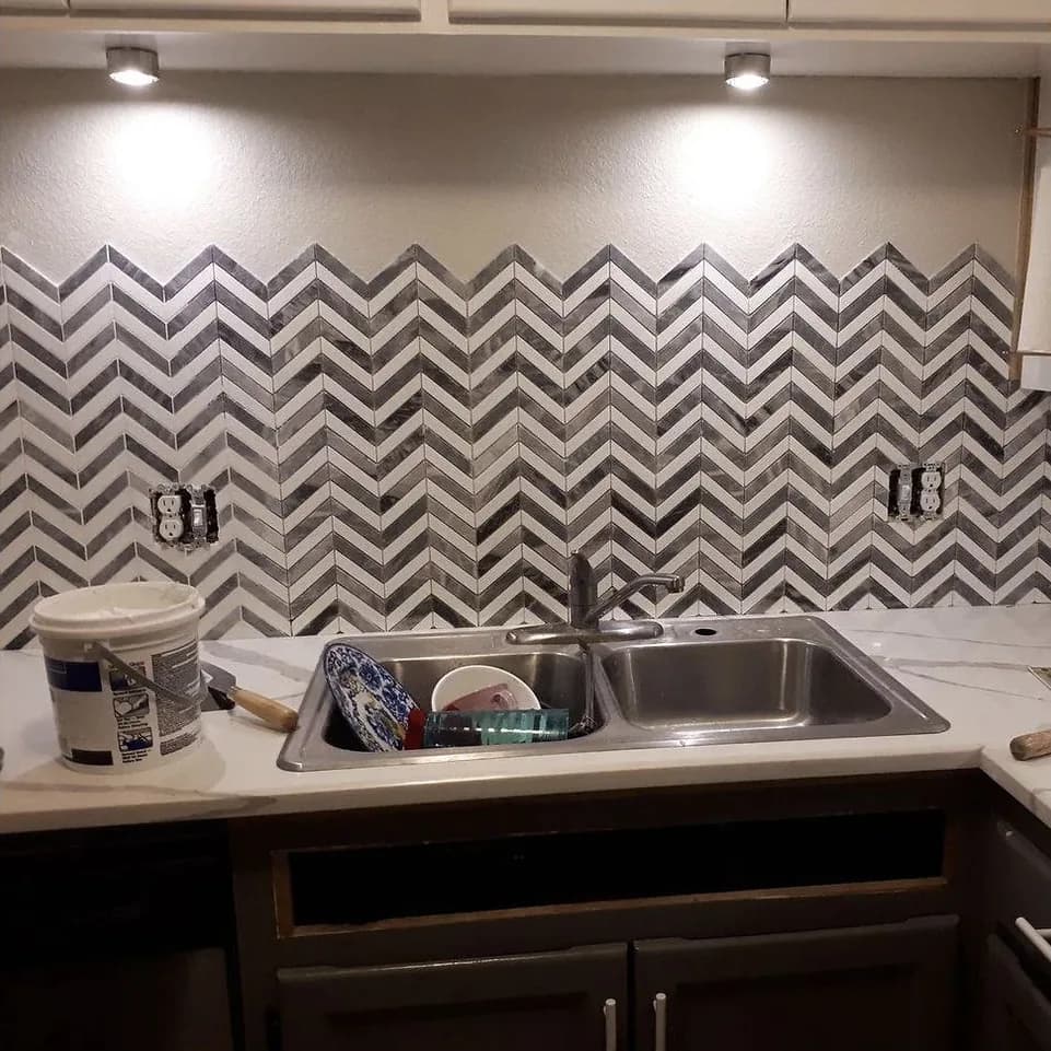 A chevron marble mosaic backsplash under cabinet lights, mid-install, with outlet covers off and dishes in the sink
