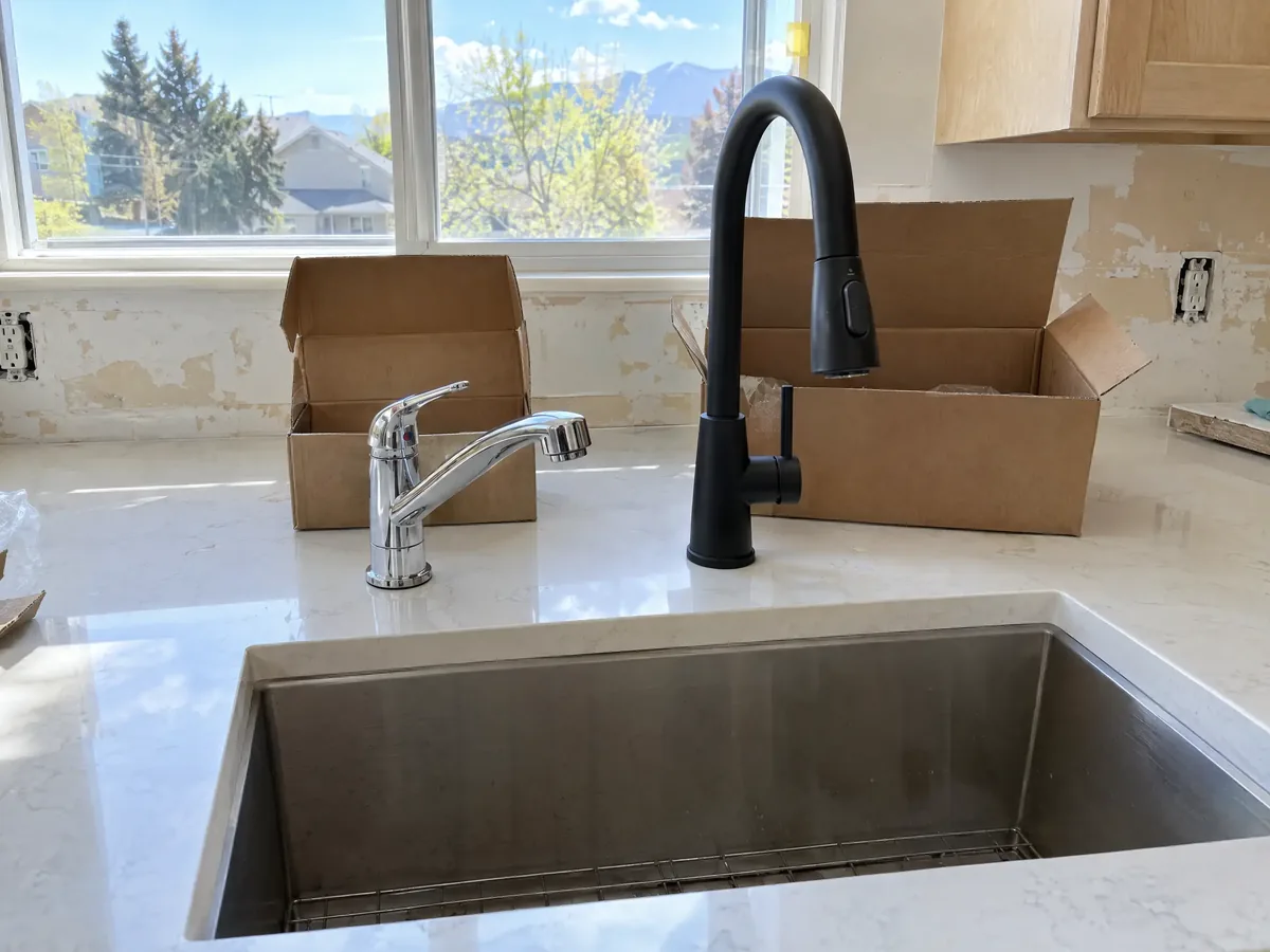 A plain chrome kitchen faucet beside a tall matte black pull-down faucet on a new quartz counter