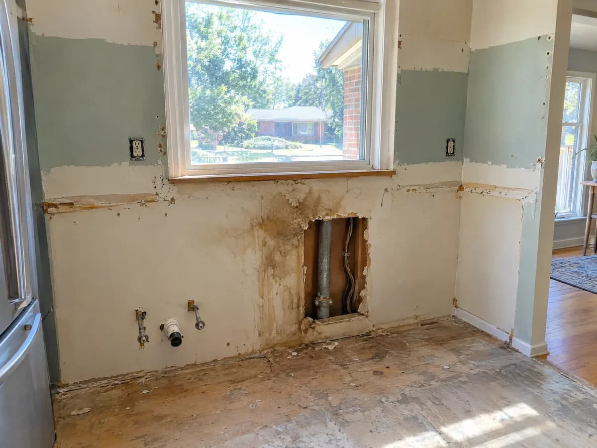 A kitchen wall after the cabinets came off, with an old water stain, an old pipe and old wiring showing through a cut-open patch