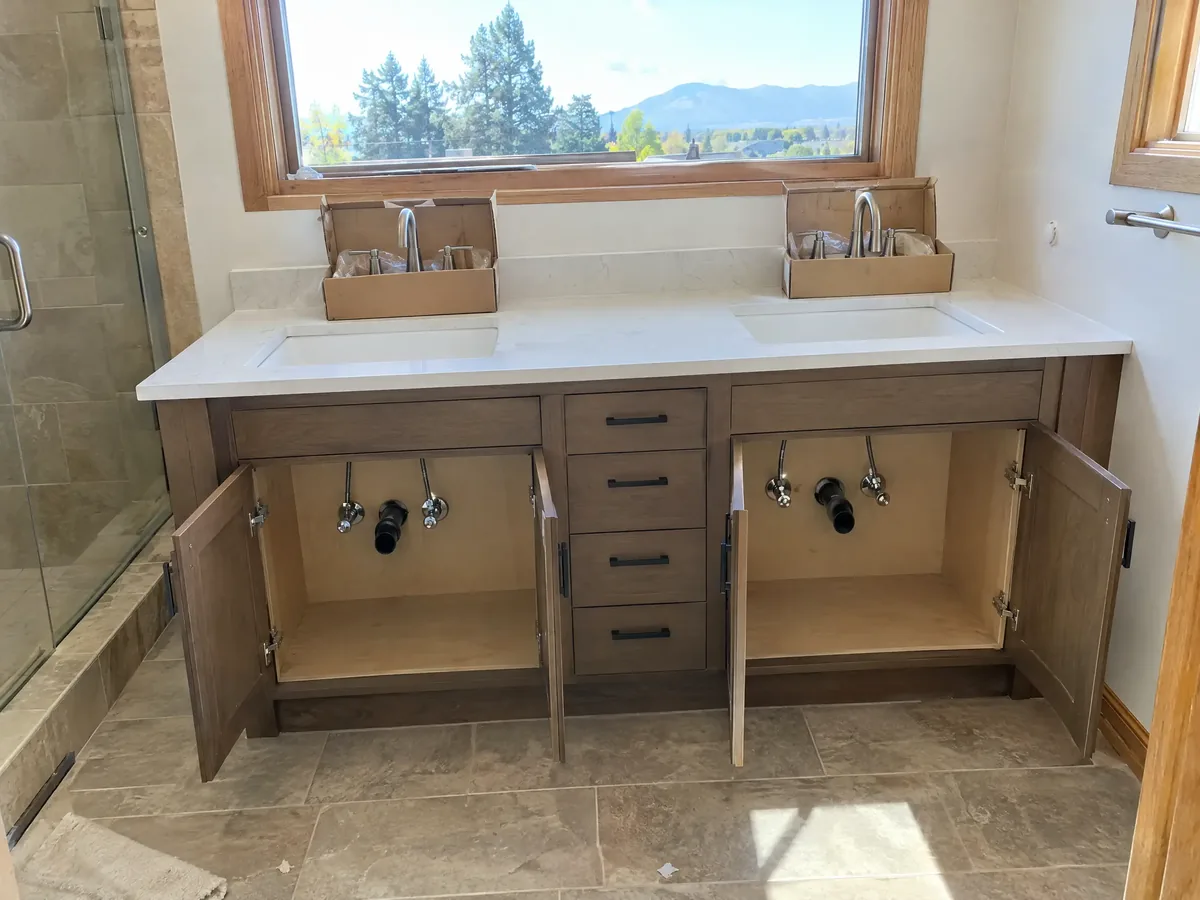 A double vanity mid-install with its doors open, showing two sets of supply valves and two drains