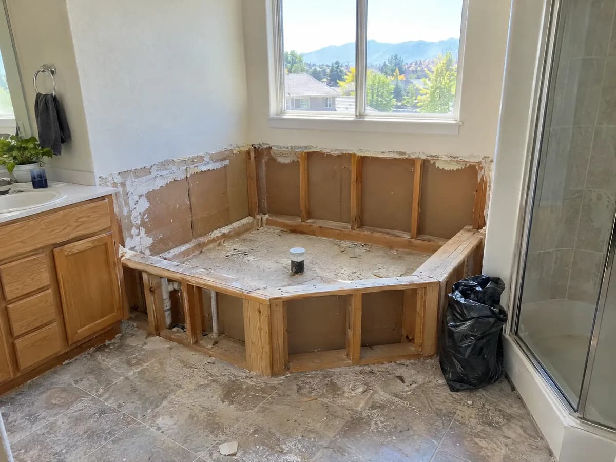 A primary bathroom mid-remodel with an old corner garden tub torn out, leaving its framed platform and a capped drain