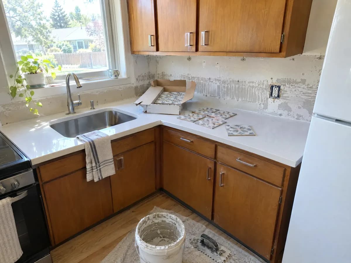 A short run of new quartz countertop with an opened box of patterned tile, a trowel and a bucket of thinset below
